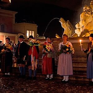 Tänzerinnen in Tracht mit Blumensträußen im Arm haben bei den Stufen des Residenzbrunnens Aufstellung genommen