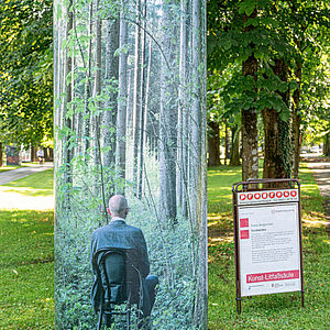 grüne Kunst-Litfaßsäule, welche fast mit der Natur verschwimmt am Ignaz-Rieder-Kai (Radweg Volksgarten)