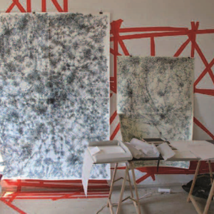 Foto des Ateliers mit zwei Bildern "Odyssee" an der Wand davor ein mit Papier bedeckter Tischm 