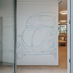 Blick durch eine geöffnete Glatür auf einen VW Käfer an der gegenüberliegenden Wand 
