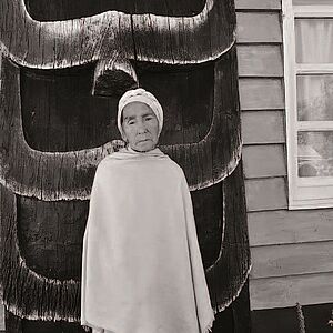 Fotografie einer alten Frau mit hellem Turban, eingehüllt in ein helles Tuch, darunter Beine in hellen Hosen, vor einer übermannshohen Art Holzstatue, die Naga (eine Schangengottheit) symolisiert