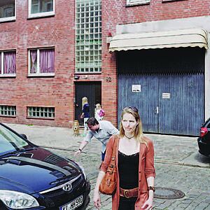 Foto einer blonden Frau mit die vor einem parkenden Auto eine gepflasterte Straße überquert.