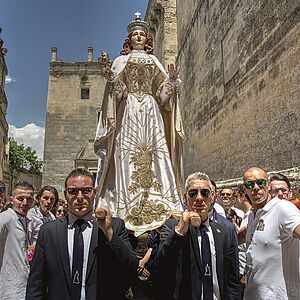 Eine lebensgroße Madonnen Statue in einem weiß-goldkordel verzirtem langen Kleid mit Umhang und einer Krone auf dem Kopf auf zwei Tragestangen, die von vier Männern im schwarzen Anzug durch die Menschenmenge getragen wird.