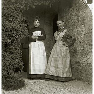 Schwarz-Weiss Fotografie von zwei Arbeiterinnen mit langen Kleiderschürzten in einem Torbogen.