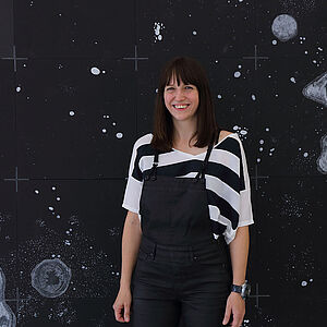 Foto von Marianne Lang vor einem Bild mit einem Sternenhimmel und größeren und kleineren Asteroiden  