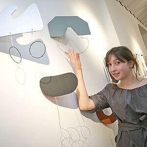 Veronika Atzwanger deutet auf ihre im feien Raum hängenden Skulpturen unterschiedlichen Formen mit daran hängenden Ringen oder Wellen aus Aluminiumdraht