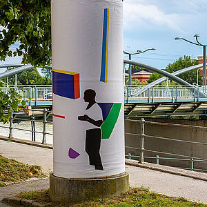 Litfaßsäule mit dem Kunstplakat; im Hintergrund die Karolinger Brücke mit dem Künstlerhaus am gegenüberliegenden Salzachufer