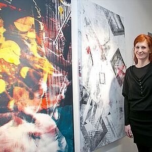 Martina Stock steht neben ihren mannshohen Werken - zwei abstrakte Bilder auf Leinwand, eines in gelb-rot gehalten eines in bläulich-weiß.