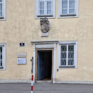 Frontale Ansicht der Eingangsseite des Gebäudes mit der geöffneten Eingangstür, links daneben hängt das Schild für die Bewohnerservice-Stelle