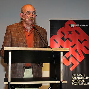 Der Vortragender Gert Kerschbaumer steht auf einem Podest und haelt einen Vortrag zum Thema Nationalsozialismus wo er Dinge wie Schicksaele von Kriegsdienstverweigerern, Homosexuellen, Kindern und vieles mehr anspricht.