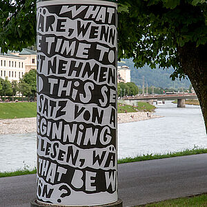 Litfaßsäule mit dem Kunstplakat im Hintergrund der Geh- und Radweg entlang der Salzach und der Fluss