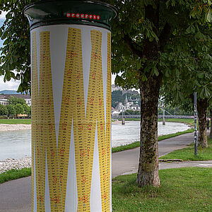 Litfaßsäule mit dem Kunstplakat, im Hintergrund der Gehweg entlang der Salzach und der Fluss