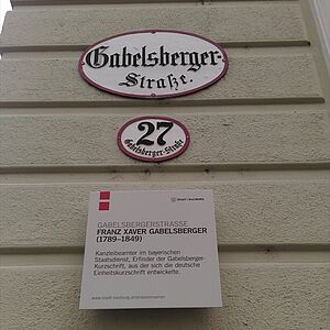 Foto-Ausschnitt der Hausfassade mit Strassennahmenschild zuoberst, darunter das Hausnummernschild und darunter die Erlaeuterungstafel.