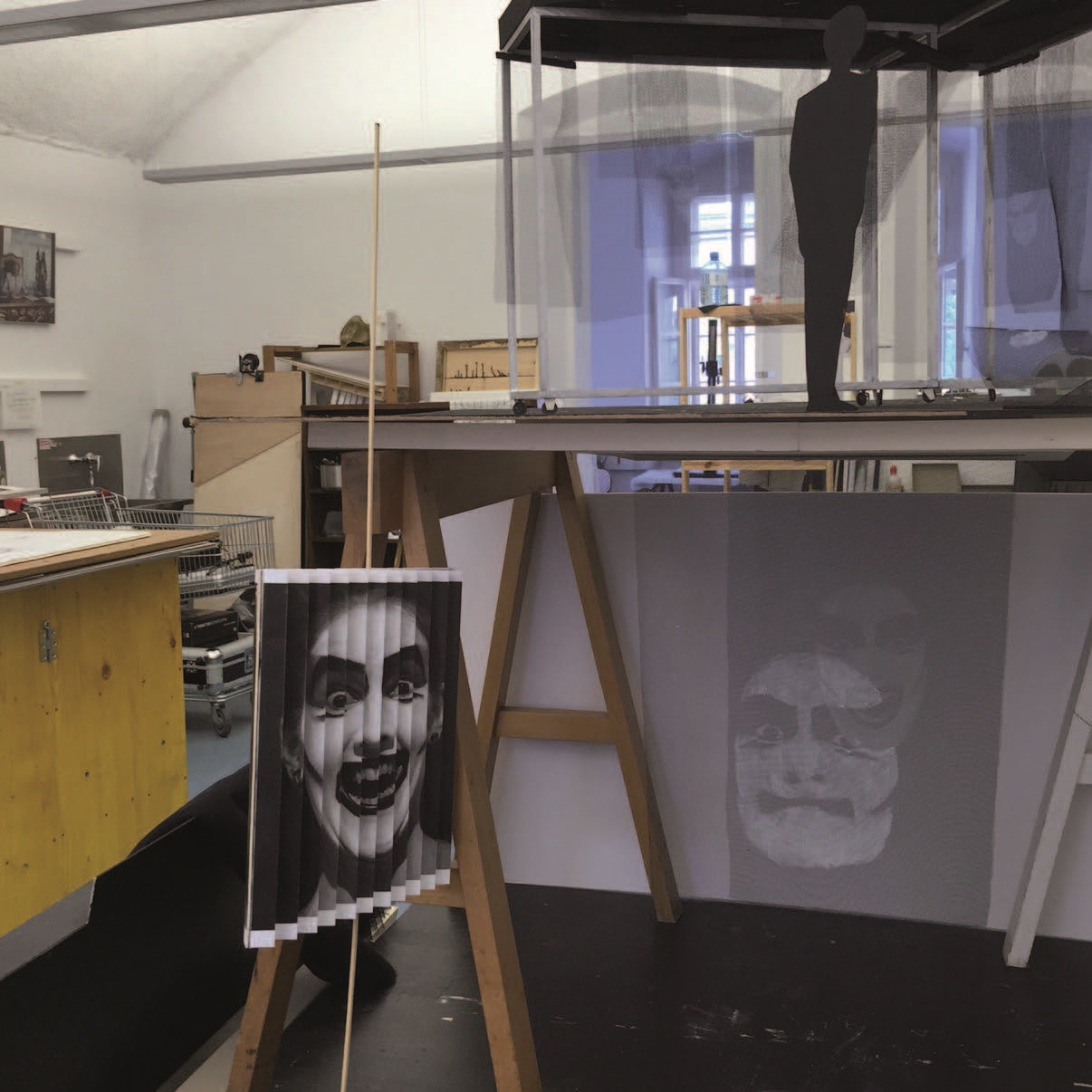 "How to become Frieda" Werkstatt mit aufgebockter hoher Tischplatte auf der eine Installation steht, davor ein Bild mit einem stark geschmickten schwarz-weißem Gesicht - gefaltet wie ein Fächer - an einem senkrechten Stab.