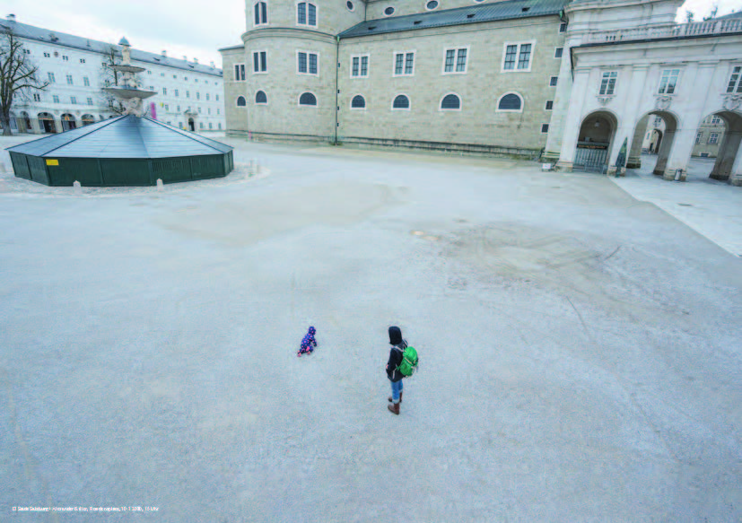 Foto vom Residenzplatz mit dem Residenzbrunnen, der mit der Winterverkleidung aus Holz abgedeckt ist. Im Vorderfgrund eine erwachsene Person mit einem spielenden Kind, sonst ist der Platz Menschenleer.
