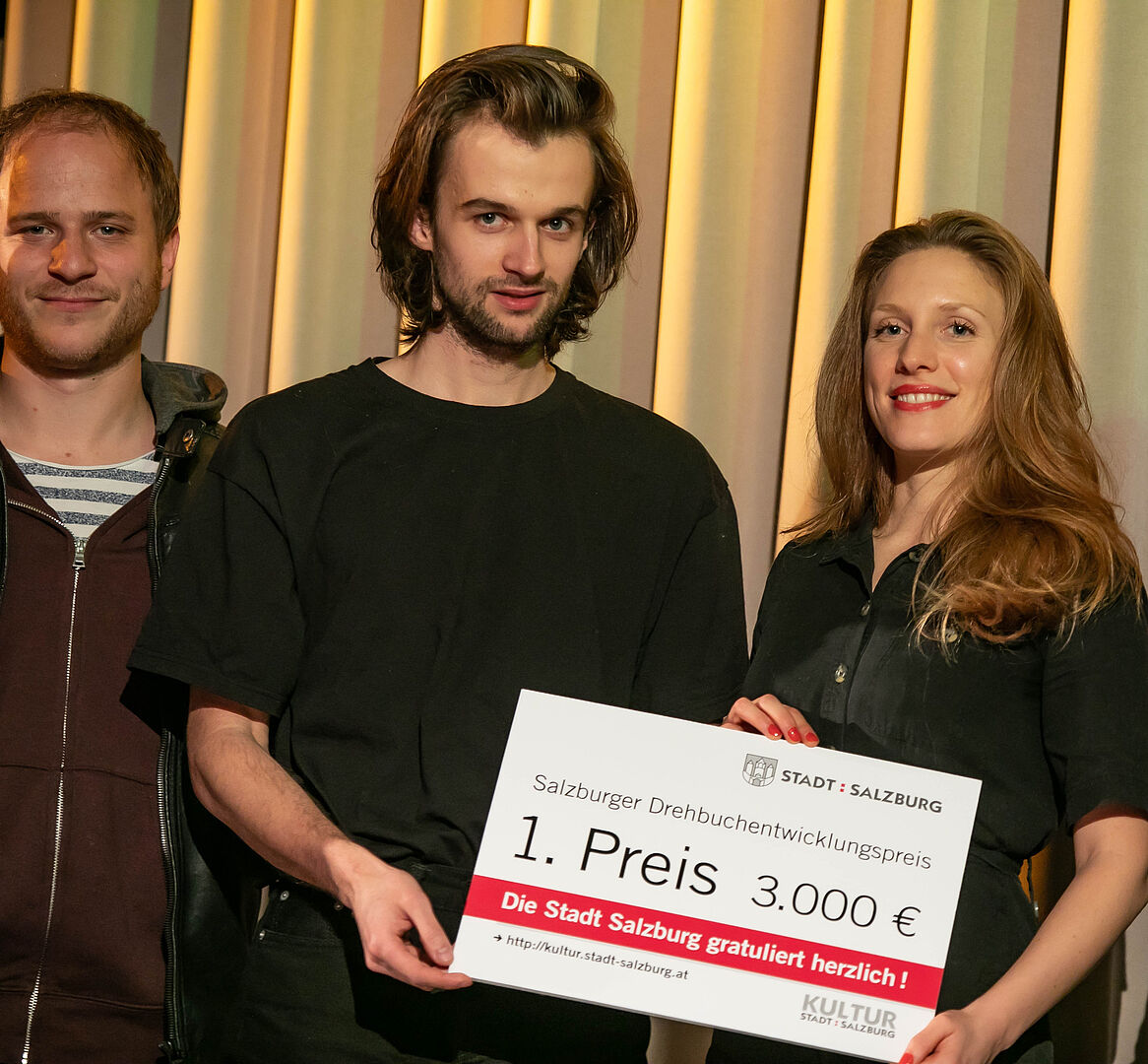 Die beiden Erst Platzierten nebeneinander, die das Hauptpreis-Schild halten, links im Hintergrund der Juror Adrian Goinger 