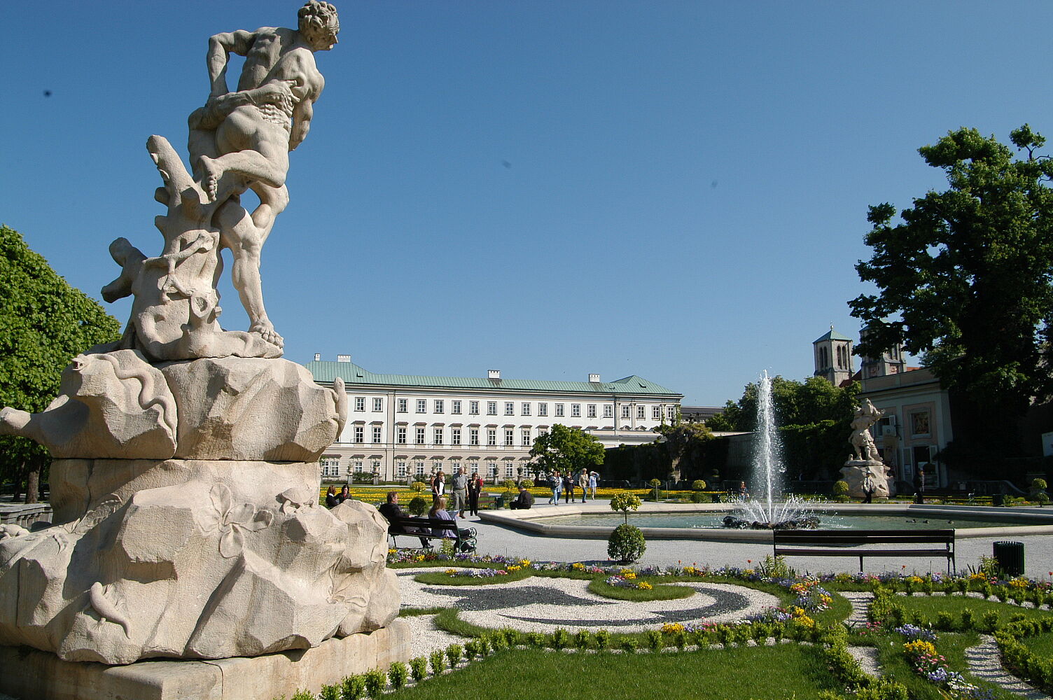 Links im Vordergrund ist eines der vier Figurenpaare zu sehen, dahinter rechts im Bild der Brunnen und im Hintergrund des Fotos das Schloss Mirabell