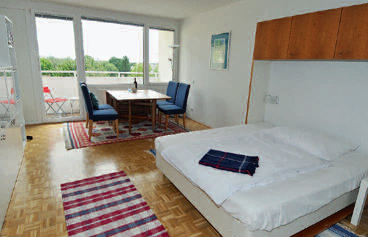 Wohnung Tobias Das Wohn- Schlafzimmer mit Blick in Richtung Balkon, rechts im Vordergrund eine zum Bett ausgeklappte Couch, dahinte vor den Raumhohen Fenstern platziert, ein Esstisch mit vier Sesseln