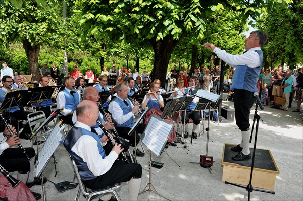 Promenadenkonzert Dirigent auf einem Podest dirigiert die vor ihm in Tracht sitzenden Musiker unter den Bäumen der Promenade im Mirabellgarten