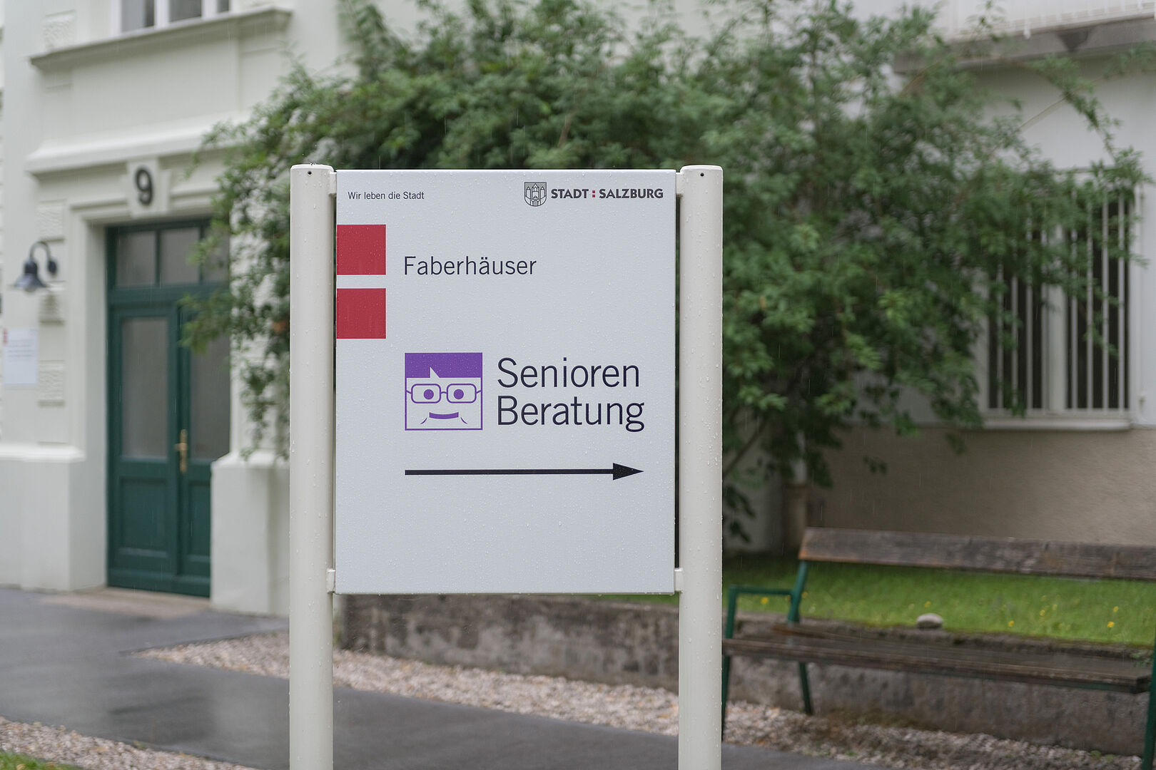 Seniorenberatung - Hinweisschild im Innenhof Faberhäuser Ein weißes Hinweisschild zwischen zwei Stangen in Augenhöhe mit dem roten Doppelpunktsymbol der Stadt Salzburg (links oben) und mittig dem Schriftzug 'Faberhäuser' und darunter 'Seniorenberatung' und darunter einen schwarzen Pfeil nach rechts. Im Hintergrund verschwommen der Innenhof der Faberhäuser.