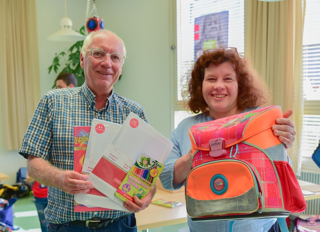 Ein Herr mit Schulheften und eine Frau mit einem Schulrucksack in den Händen, nebeneinander stehend