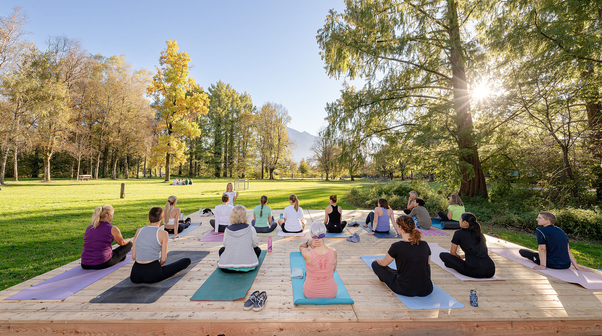 Das Bild zeigt Menschen im Park, die auf Turnmatten sitzen.