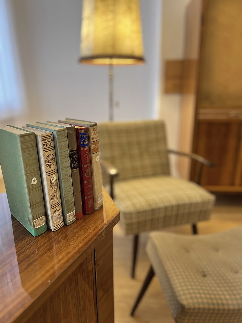 Escape Room Auf dem Bild sieht man einen ruhigen Raum. Vorne stehen 6 Bücher auf einem Schrank aus Holz. Die Bücher haben bunte Buch-Rücken. Man kann die Titel auf den Büchern lesen. Hinter dem Schrank steht ein weicher Sessel. Der Sessel ist kariert und hat Armlehnen. Davor steht ein Hocker im gleichen Muster. Im Hintergrund sieht man eine hohe Lampe. Die Lampe macht warmes Licht. Der Raum sieht gemütlich aus.
